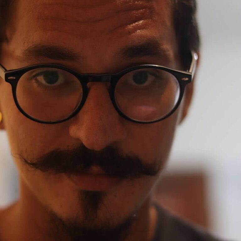 Homem com óculos e bigode olhando para a você em um enquadramento que o faz parecer muito perto